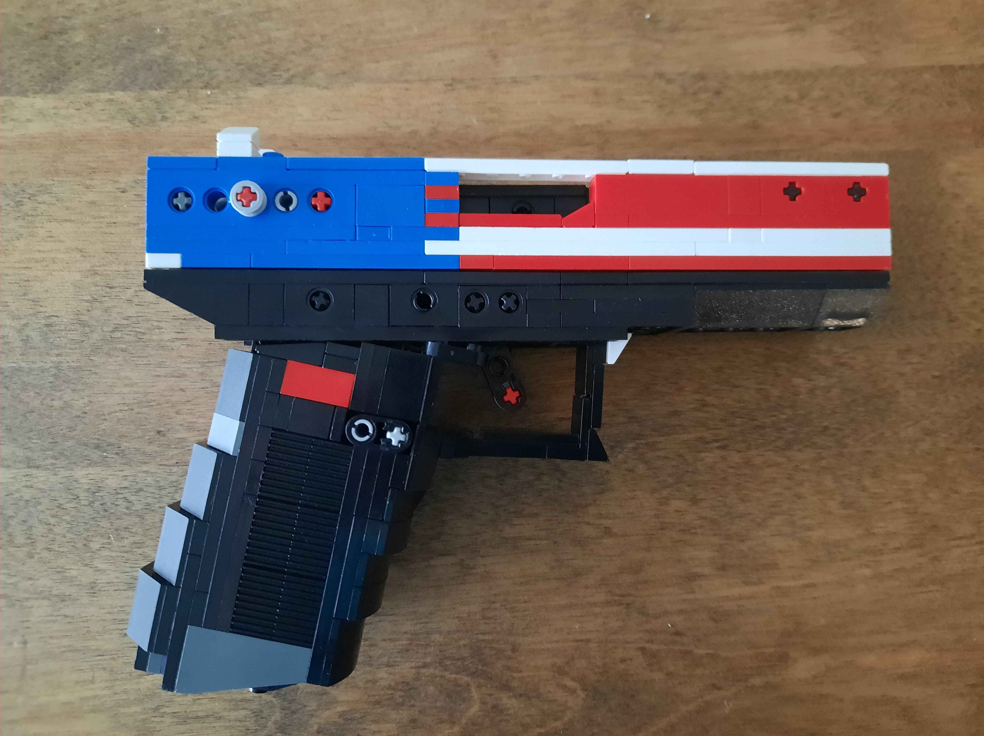 A custom LEGO model of a handgun, featuring a combination of black, blue, white, and red colors, displayed on a wooden surface.